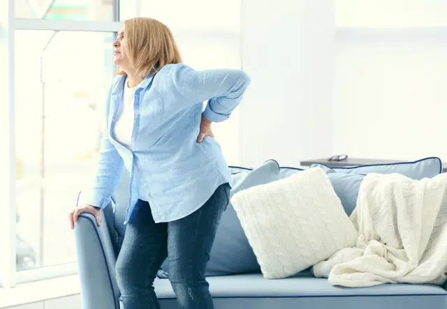 elder woman holding her back due to pain clenching the arm of the couch